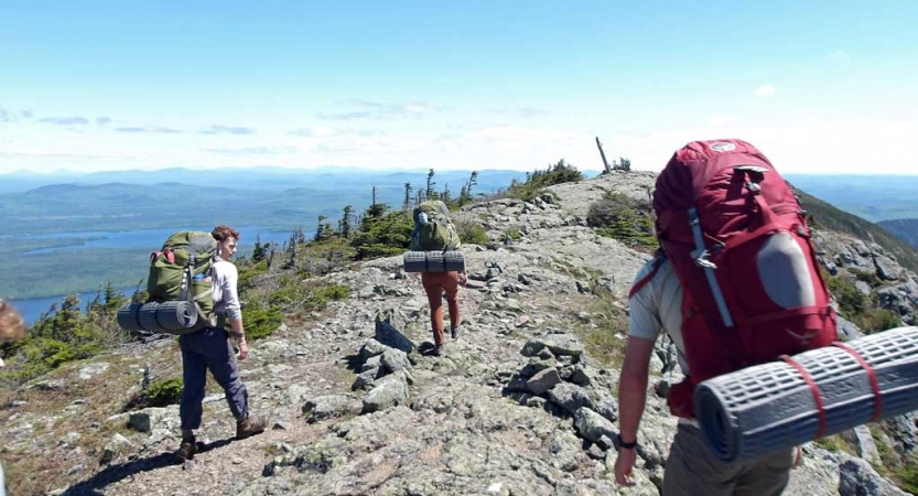 Three people wearing backpacks approach a rocky summit under blue skies